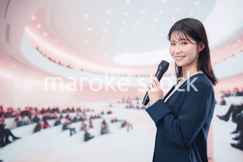 ホールで司会・プレゼンをするスーツを着た若い女性