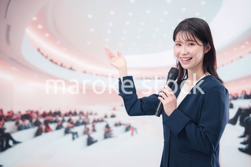 ホールで司会・プレゼンをするスーツを着た若い女性
