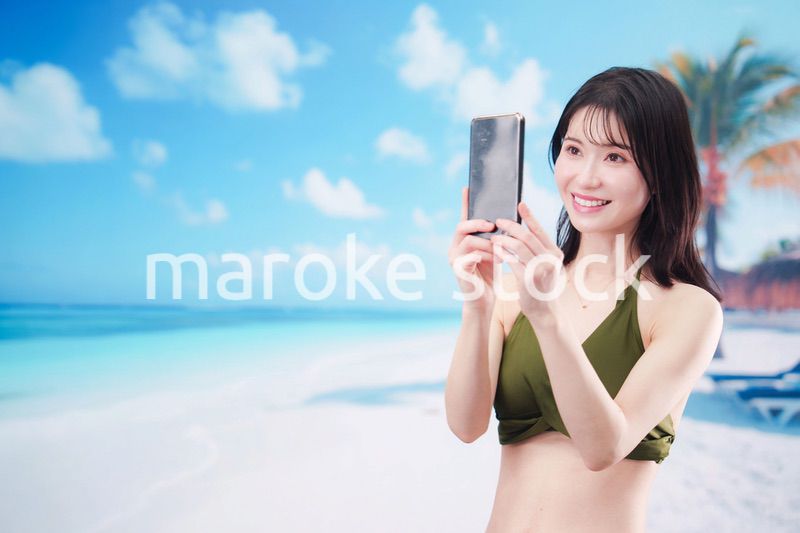 快晴のビーチでスマホで自撮りをする水着を着た若い女性