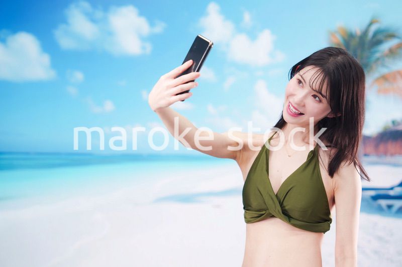 快晴のビーチでスマホで自撮りをする水着を着た若い女性