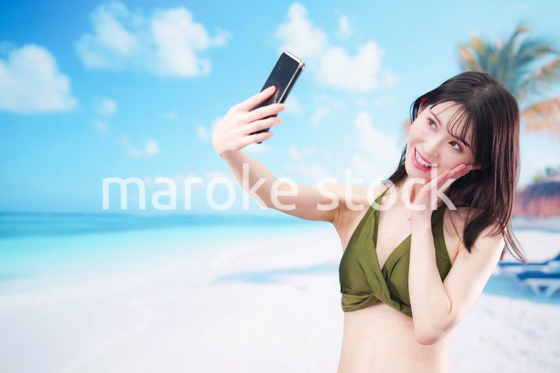 快晴のビーチでスマホで自撮りをする水着を着た若い女性