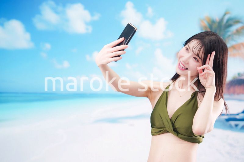 快晴のビーチでスマホで自撮りをする水着を着た若い女性