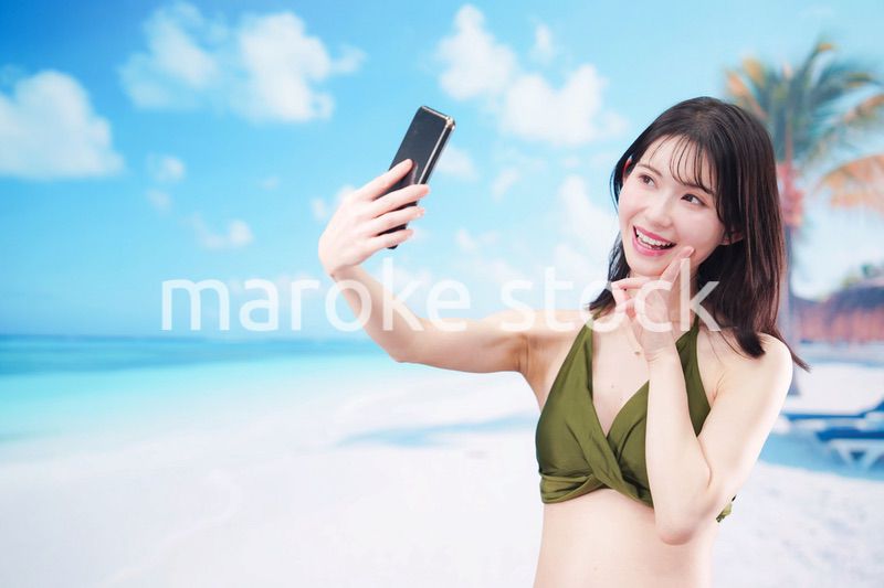 快晴のビーチでスマホで自撮りをする水着を着た若い女性