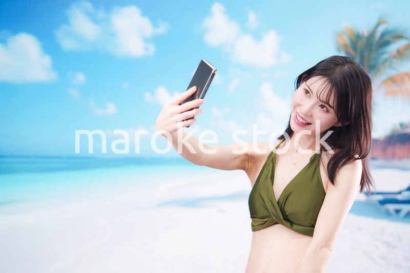 快晴のビーチでスマホで自撮りをする水着を着た若い女性